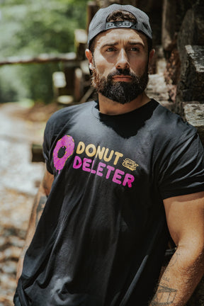 A bearded man in a backward gray cap wears a Tango Charlie Apparel Donut Deleter Tee with a donut graphic, standing by stacked logs outdoors as sunlight filters through the trees, highlighting his soft cotton-poly blend shirt.