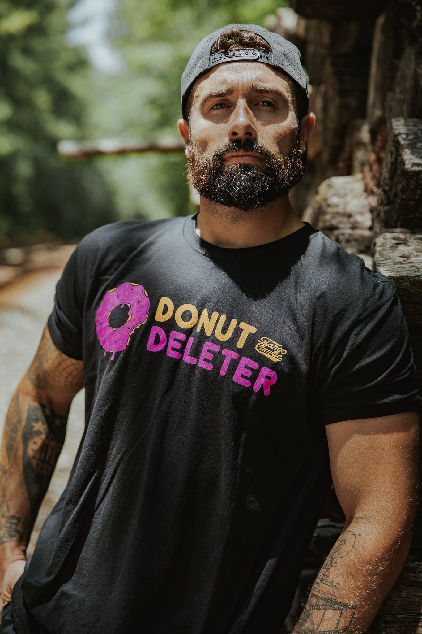 A bearded man in a backwards cap stands outdoors by stacked logs, wearing the Tango Charlie Apparel Donut Deleter Tee—a black cotton poly blend shirt featuring "DONUT DELETER" text and a bitten pink donut graphic.