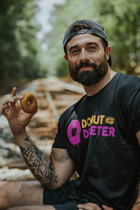 A bearded man in a backward cap and Tango Charlie Apparel Donut Deleter Tee, made from a cotton-poly blend, holds a donut while sitting on railroad tracks in a wooded area, smiling at the camera.