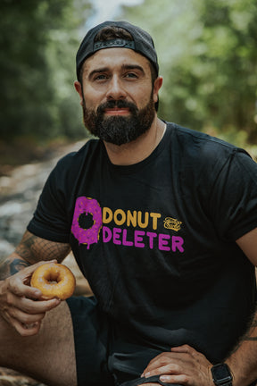A bearded man in a backward cap sits by a stream in the Tango Charlie Apparel Donut Deleter Tee, a soft black cotton-poly shirt with a pink donut graphic, standing out against the lush green woods.