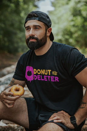 A bearded man in a backward cap sits outdoors, holding a donut. He wears Tango Charlie Apparel's Donut Deleter Tee, featuring colorful “DONUT DELETER” lettering, with blurred trees and greenery in the background.