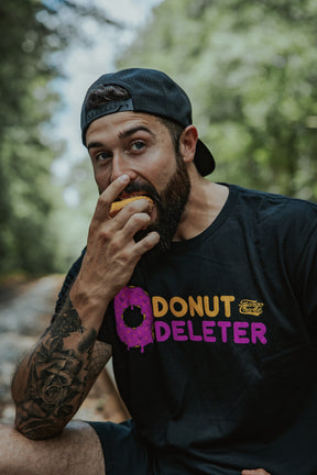 A bearded man in a backward cap enjoys a donut outdoors while wearing the Donut Deleter Tee from Tango Charlie Apparel, with trees and a blurred path in the background.