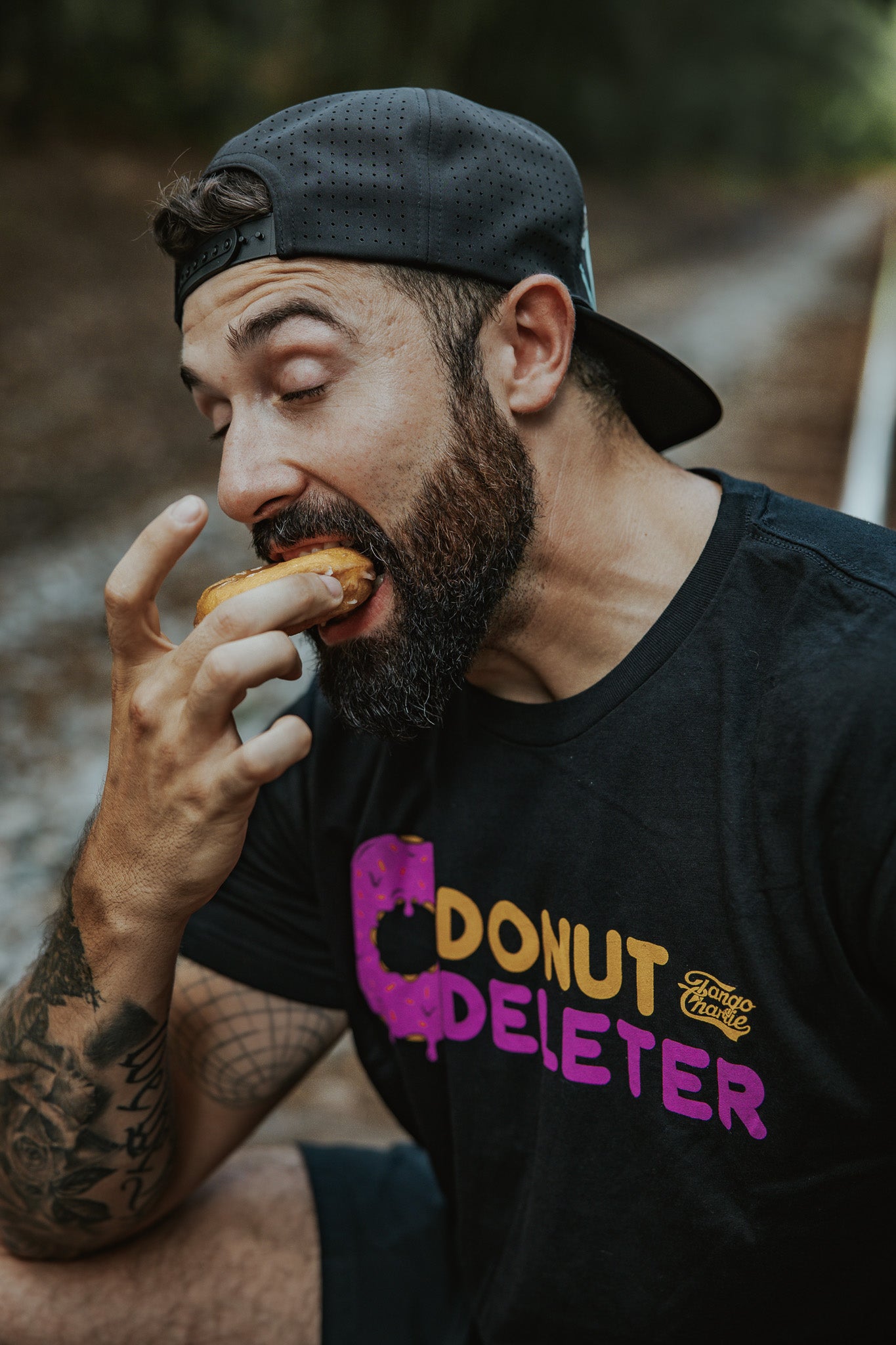 A bearded man in a black cap, eyes closed, enjoys a donut outdoors while wearing the Tango Charlie Apparel Donut Deleter Tee, made from a soft cotton-poly blend, savoring the moment.