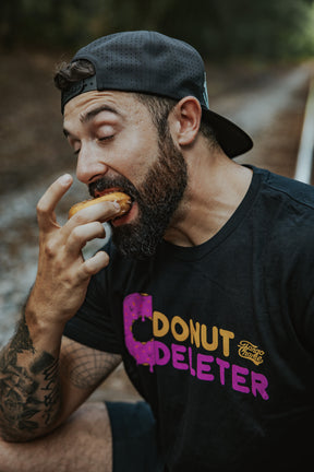 A bearded man in a black cap, eyes closed, enjoys a donut outdoors while wearing the Tango Charlie Apparel Donut Deleter Tee, made from a soft cotton-poly blend, savoring the moment.