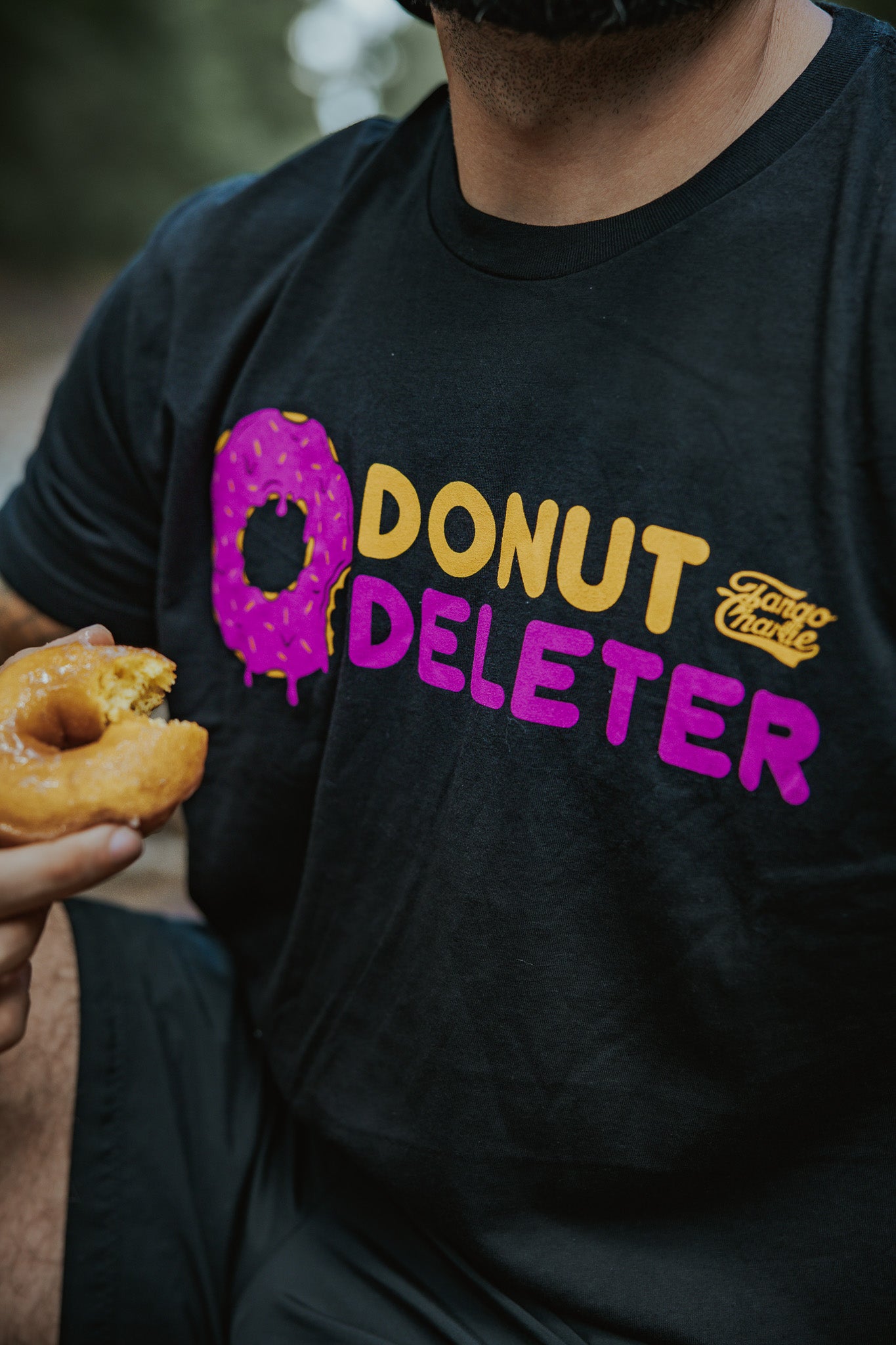 A person sports the Tango Charlie Apparel Donut Deleter Tee, a comfy black cotton-poly blend featuring "DONUT DELETER" with the O as a sprinkled donut—perfect attire for savoring treats, just like the half-eaten donut in their hand.