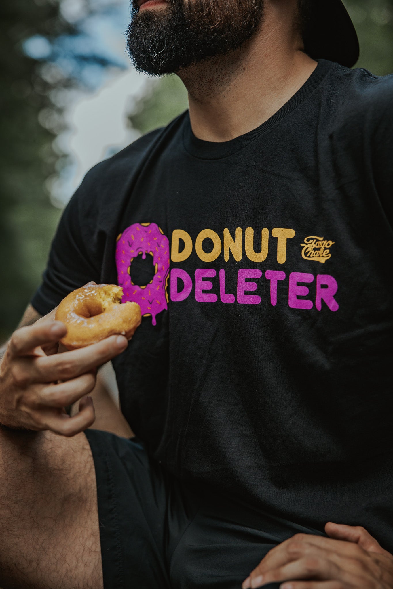 A bearded person holds a bitten donut and wears the Tango Charlie Apparel Donut Deleter Tee, which features "DONUT DELETER" with a pink frosted donut as the O, crafted from a soft cotton poly blend.