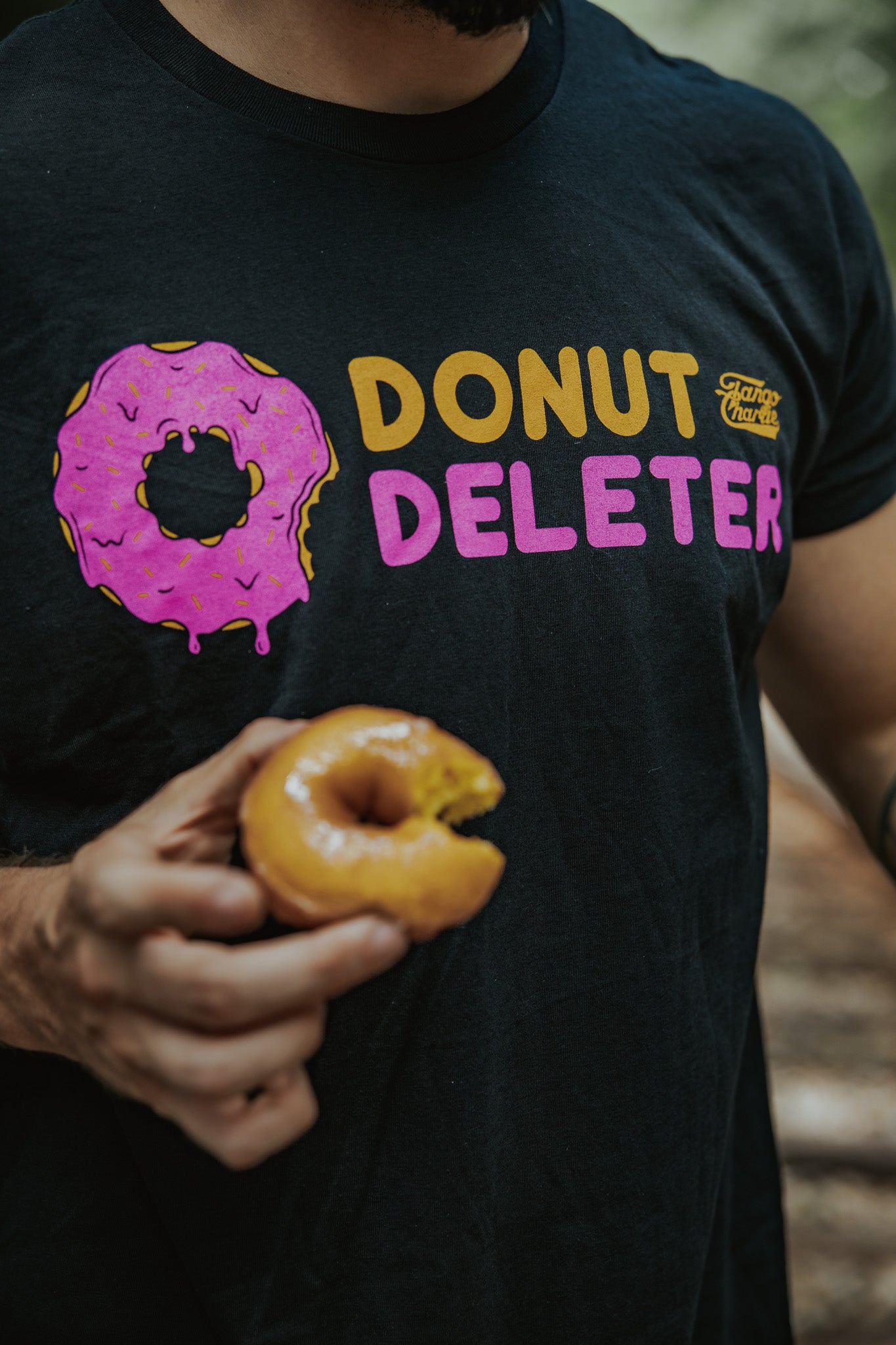 Someone wears the Tango Charlie Apparel Donut Deleter Tee—a comfy black shirt featuring a pink frosted donut graphic—while holding a bitten glazed donut.