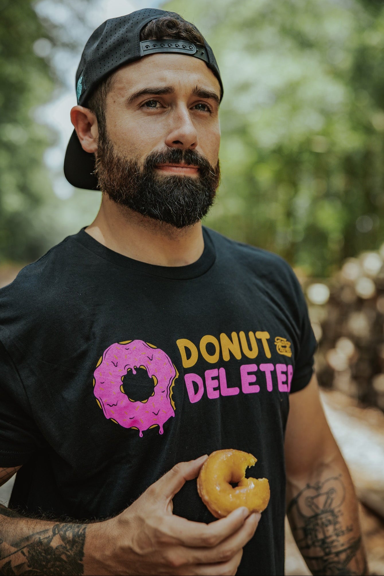 A bearded man in a backward cap and a black Tango Charlie Apparel Donut Deleter Tee holds a bitten donut outdoors, enjoying the soft cotton-poly blend of his shirt against a backdrop of blurred trees.