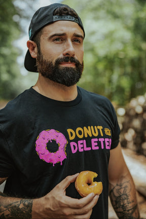 A bearded man in a backward cap and a black Tango Charlie Apparel Donut Deleter Tee holds a bitten donut outdoors, enjoying the soft cotton-poly blend of his shirt against a backdrop of blurred trees.
