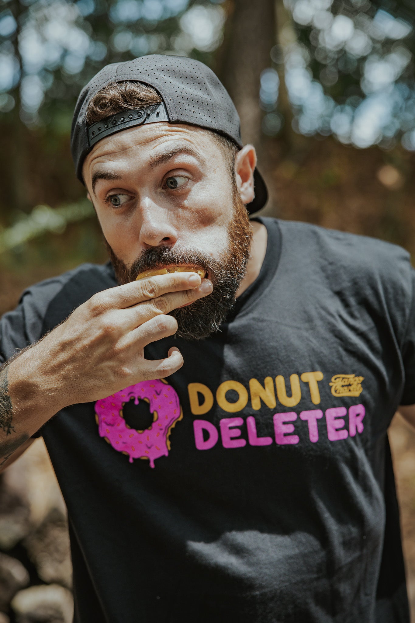A bearded man in a black backward cap wears the Tango Charlie Apparel Donut Deleter Tee, made from a soft cotton poly blend, standing outside with a playful look and his hand to his mouth as if finishing a donut.