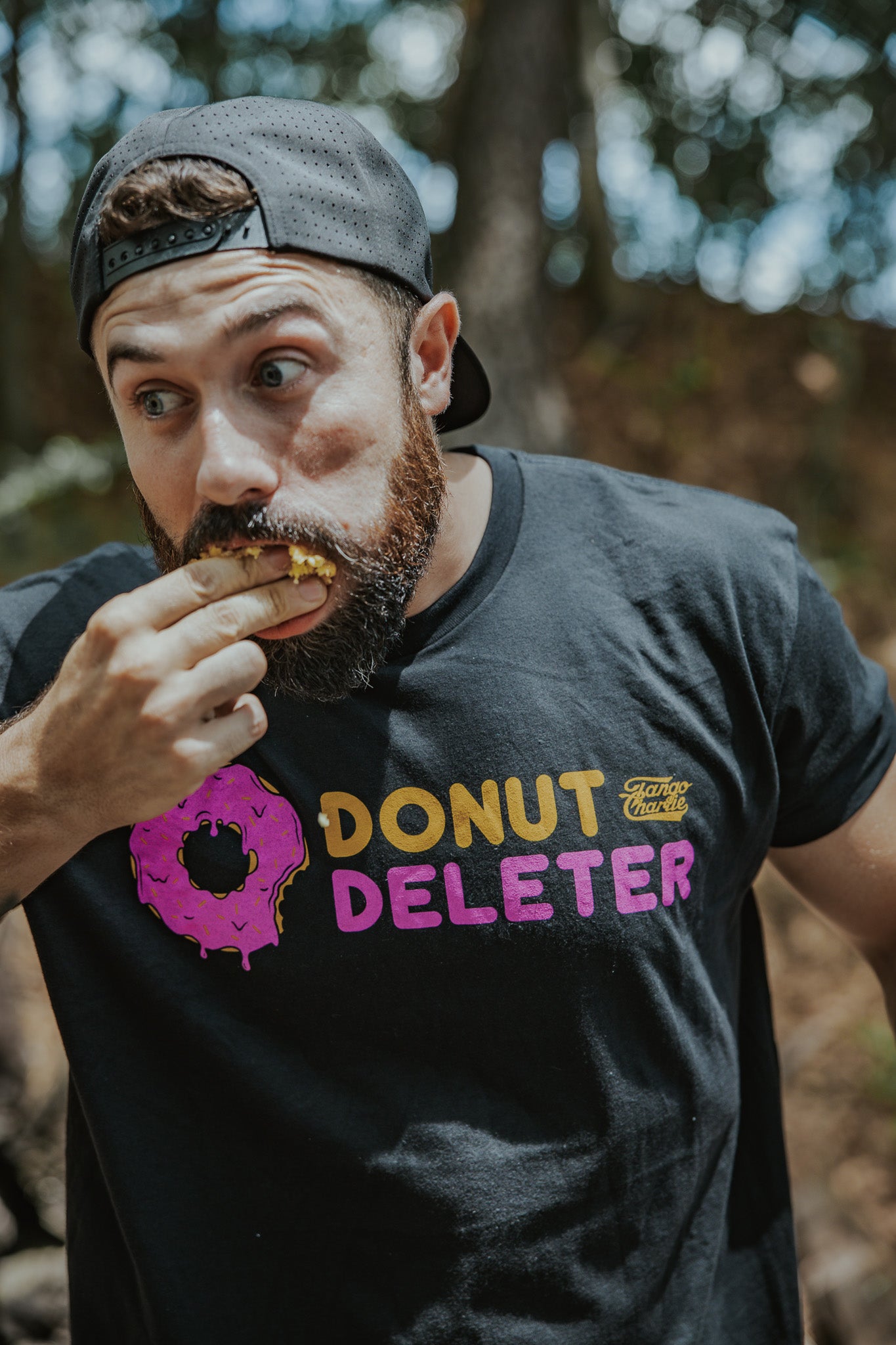 A bearded man in a backwards cap and a black Donut Deleter Tee by Tango Charlie Apparel enjoys a donut outdoors, glancing to the side with wide eyes as sunlight filters through trees—a true Tango Charlie moment.