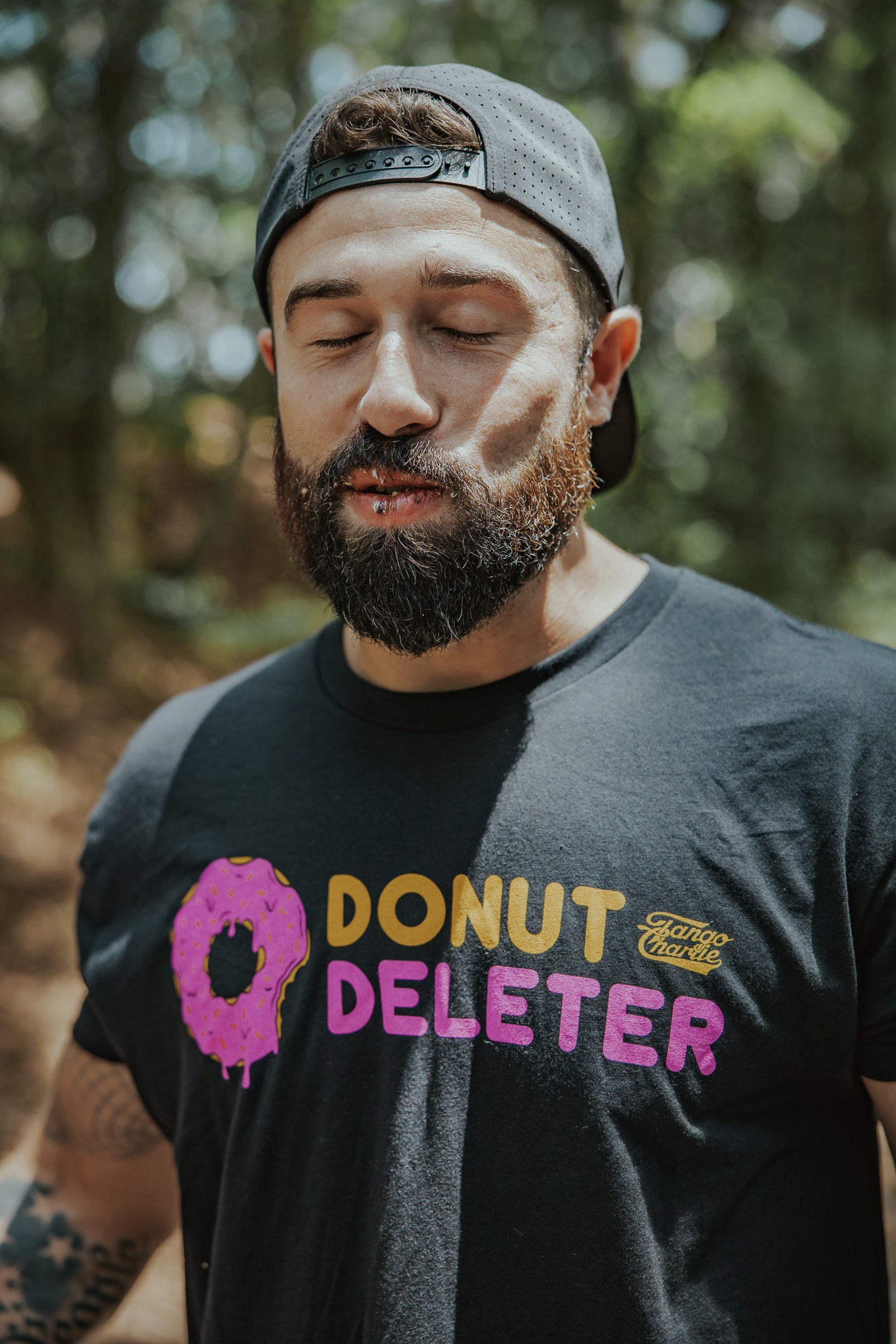 A bearded man in a black Donut Deleter Tee by Tango Charlie Apparel, featuring a pink frosted donut graphic, stands outdoors with eyes closed and a backwards cap. This cotton poly blend shirt is perfect for chill vibes.