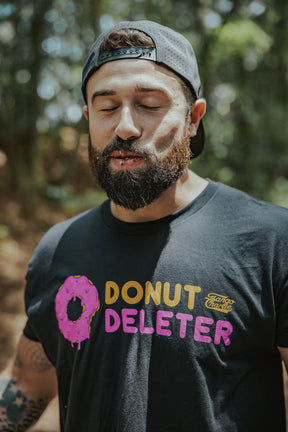 A bearded man in a black Donut Deleter Tee by Tango Charlie Apparel, featuring a pink frosted donut graphic, stands outdoors with eyes closed and a backwards cap. This cotton poly blend shirt is perfect for chill vibes.
