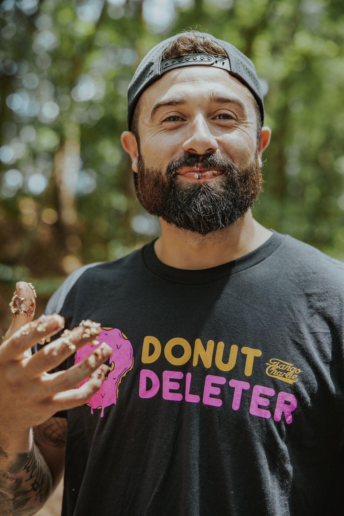 A bearded man in a backwards cap smiles outdoors, holding up a chocolate-smeared hand while wearing the Tango Charlie Apparel Donut Deleter Tee, featuring a partially eaten pink donut graphic. Trees blur softly in the background.