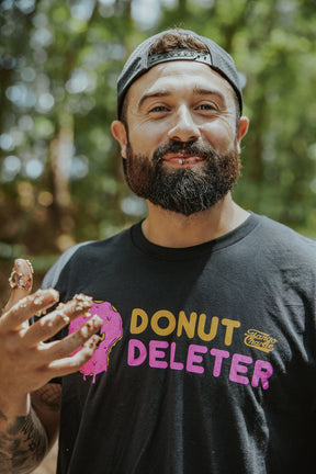 A bearded man in a backwards cap smiles outdoors, holding up a chocolate-smeared hand while wearing the Tango Charlie Apparel Donut Deleter Tee, featuring a partially eaten pink donut graphic. Trees blur softly in the background.