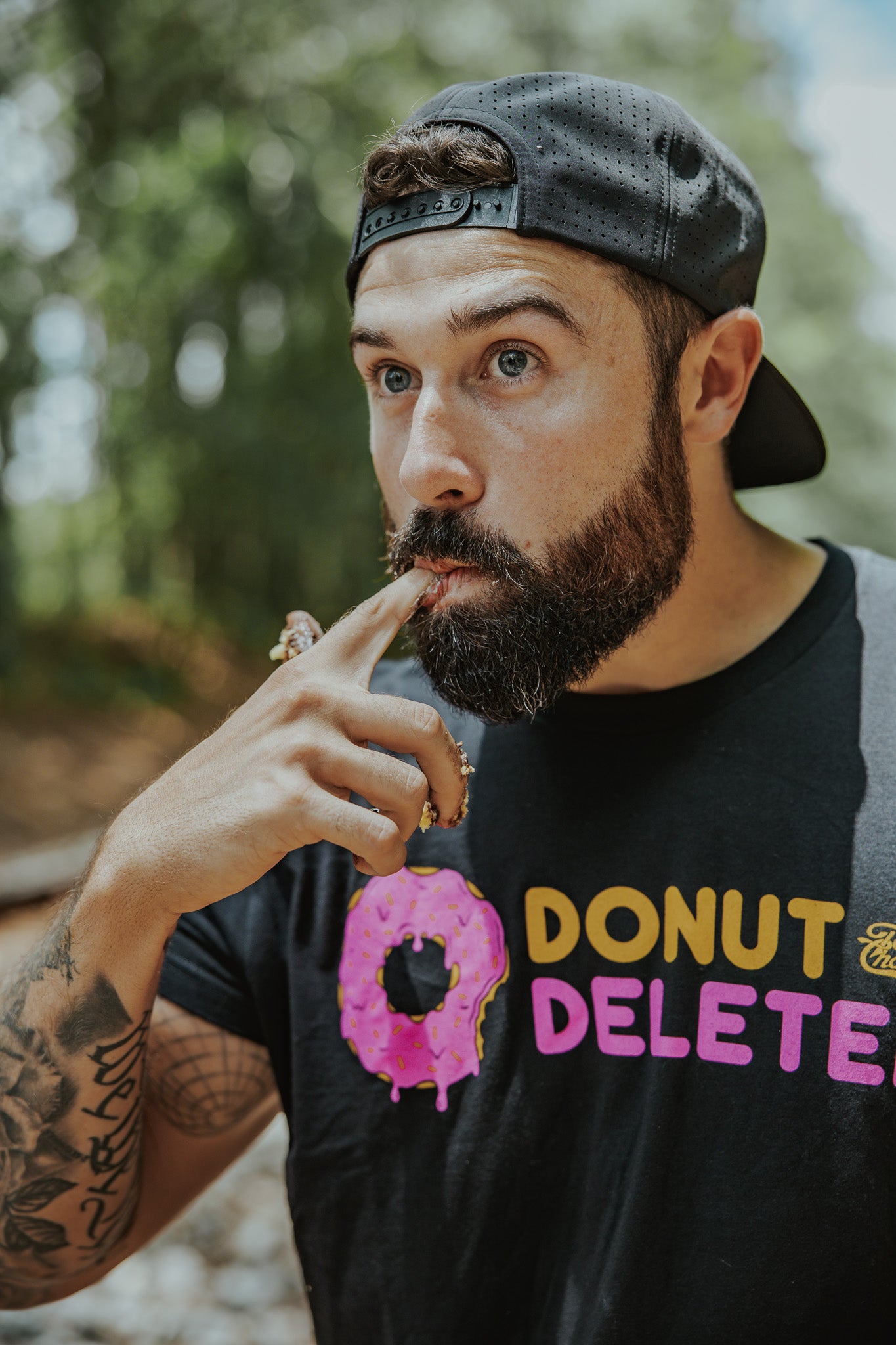 A bearded man in a backward cap and the Donut Deleter Tee by Tango Charlie Apparel licks frosting off his finger, holding a pink frosted donut. The cotton-poly blend shirt pops against the blurred background of trees and greenery.