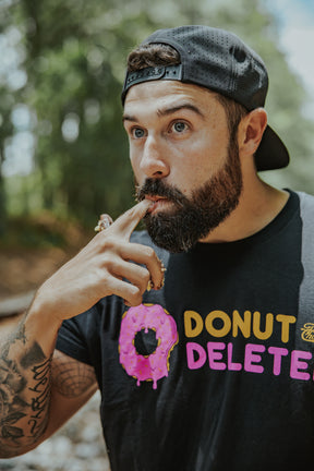 A bearded man in a backward cap and the Donut Deleter Tee by Tango Charlie Apparel licks frosting off his finger, holding a pink frosted donut. The cotton-poly blend shirt pops against the blurred background of trees and greenery.