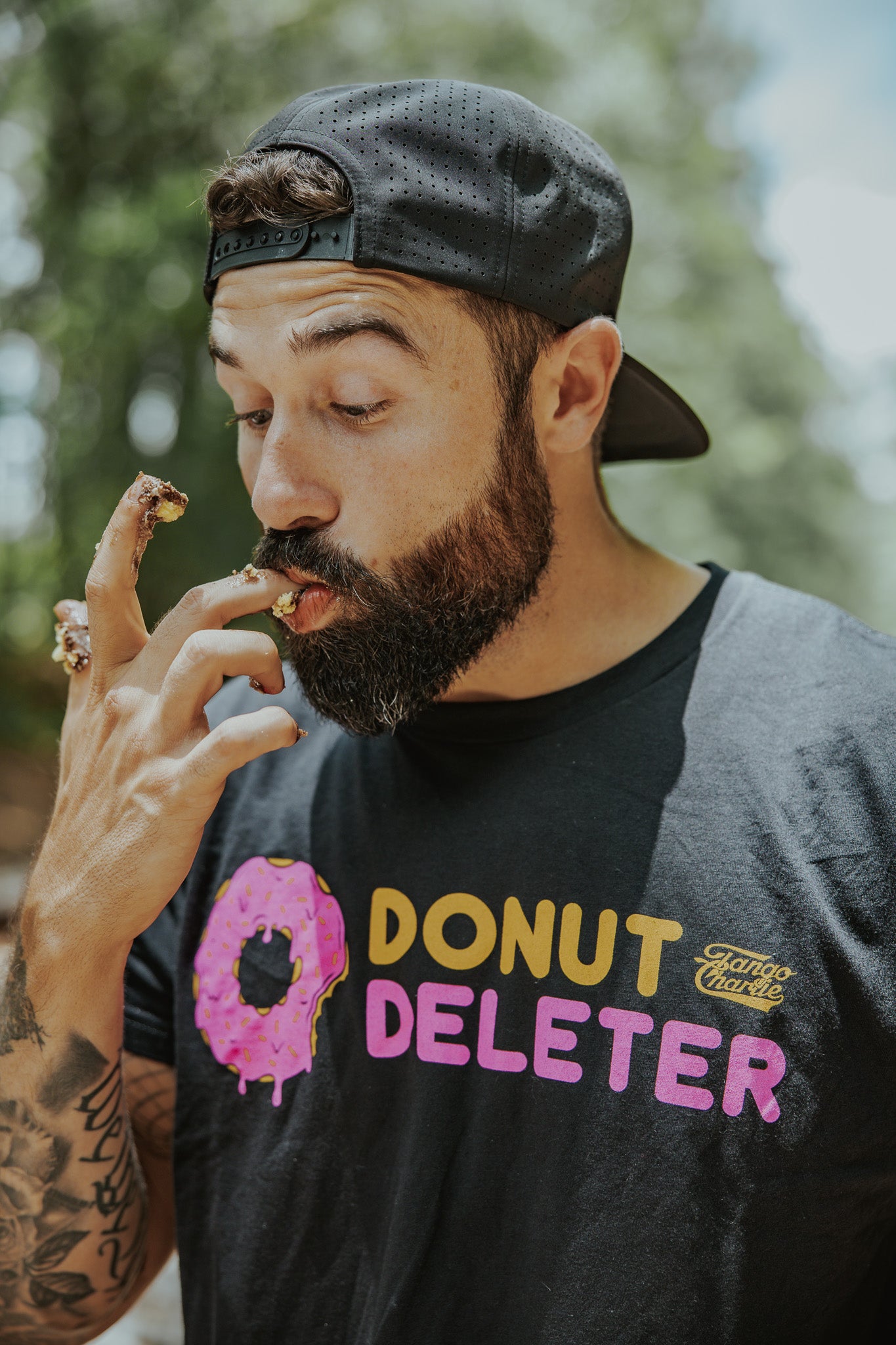 A bearded man in a black cap and Tango Charlie Apparel’s Donut Deleter Tee licks donut glaze off his fingers outdoors, with crumbs and icing visible on his comfy cotton-poly blend shirt.