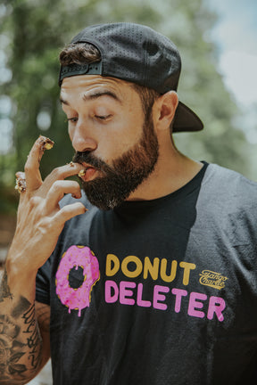 A bearded man in a black cap and Tango Charlie Apparel’s Donut Deleter Tee licks donut glaze off his fingers outdoors, with crumbs and icing visible on his comfy cotton-poly blend shirt.