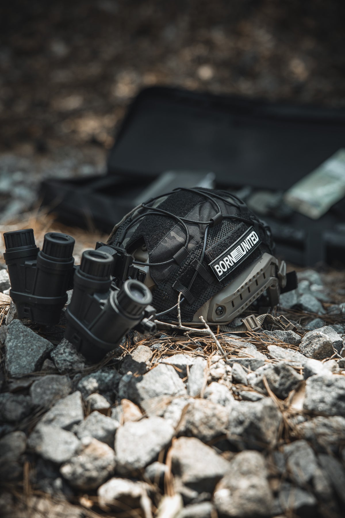 A Born United Private Label Born United PVC Patch is displayed on a tactical helmet resting on rocky ground near binoculars, with an open black case and pine needles adding to the rugged atmosphere.