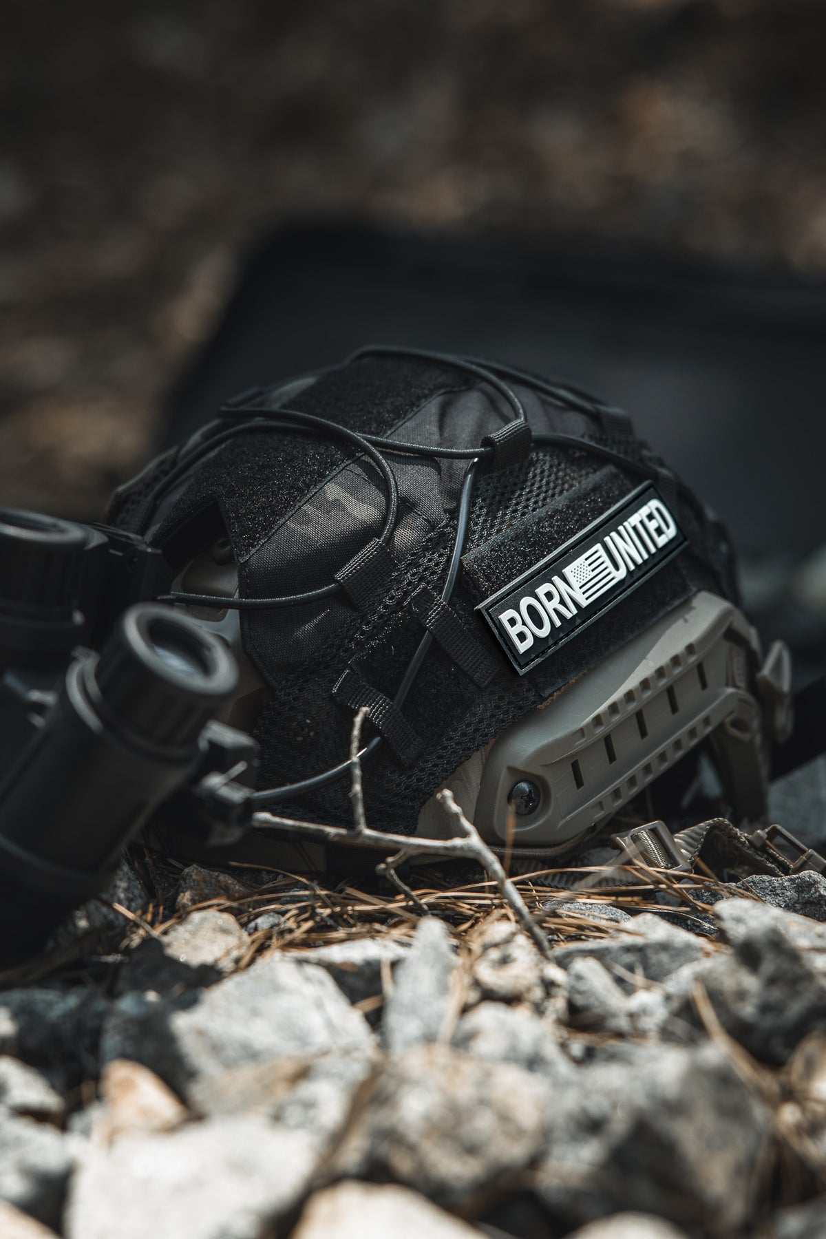 A black tactical helmet with a Born United Private Label Born United PVC Patch and night vision goggles rests on rocky ground with pine needles scattered around.