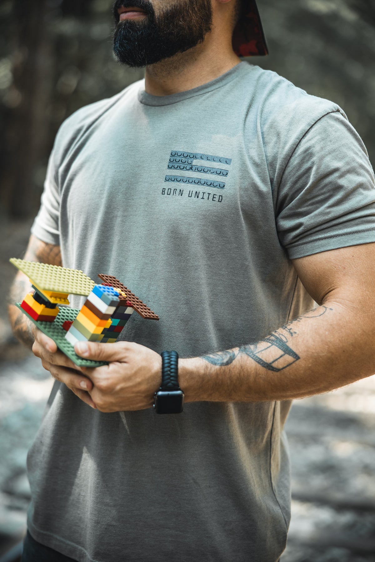 A bearded man in a gray Born United Private Label Brick Operator Tee and smartwatch stands outdoors, holding colorful LEGO bricks. His tattooed arm is visible with a blurred forest behind him.