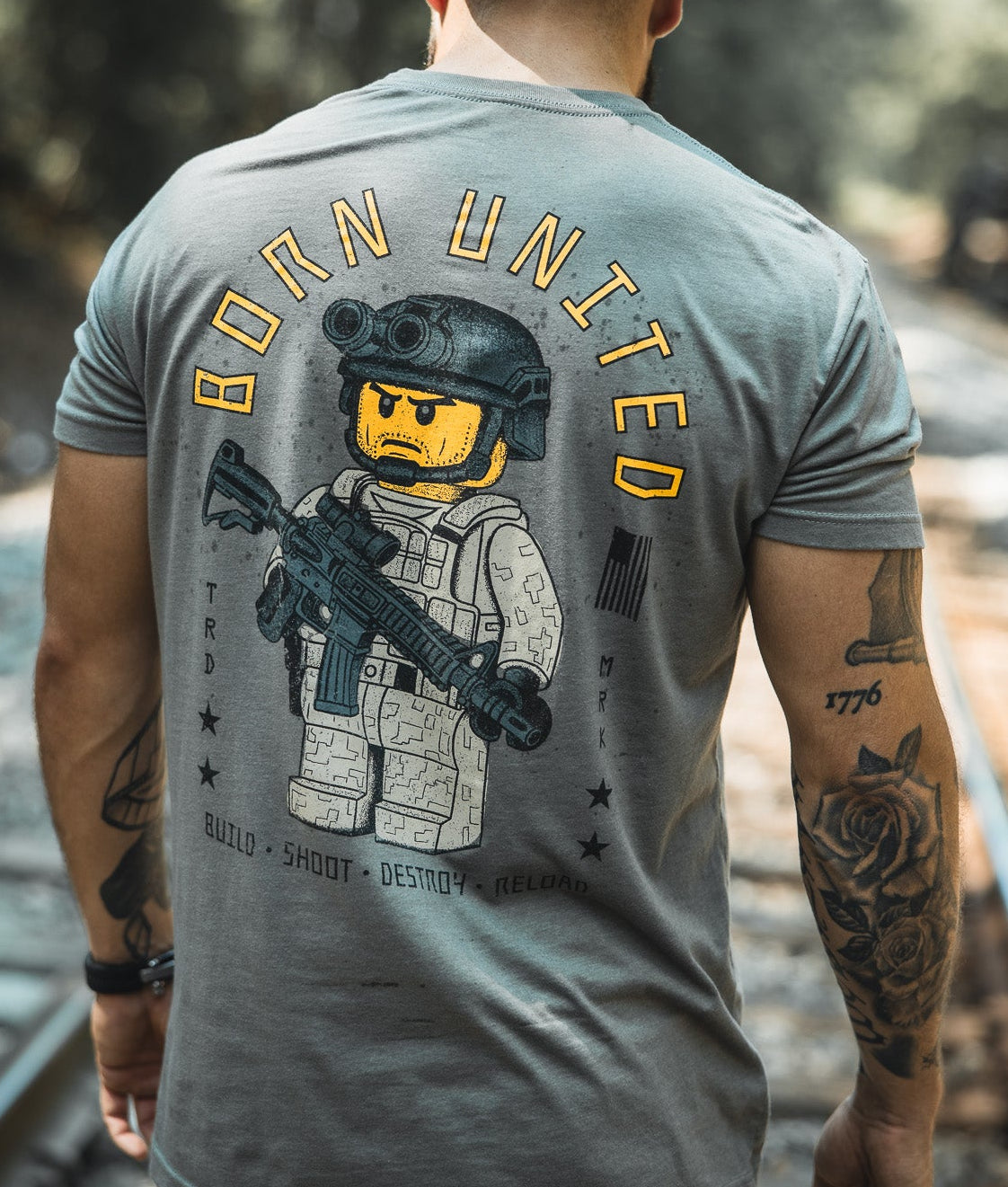 A tattooed man stands on railroad tracks in the Born United Private Label Brick Operator Tee, featuring bold yellow lettering and block-style operator art. He completes his look with a black American flag cap.
