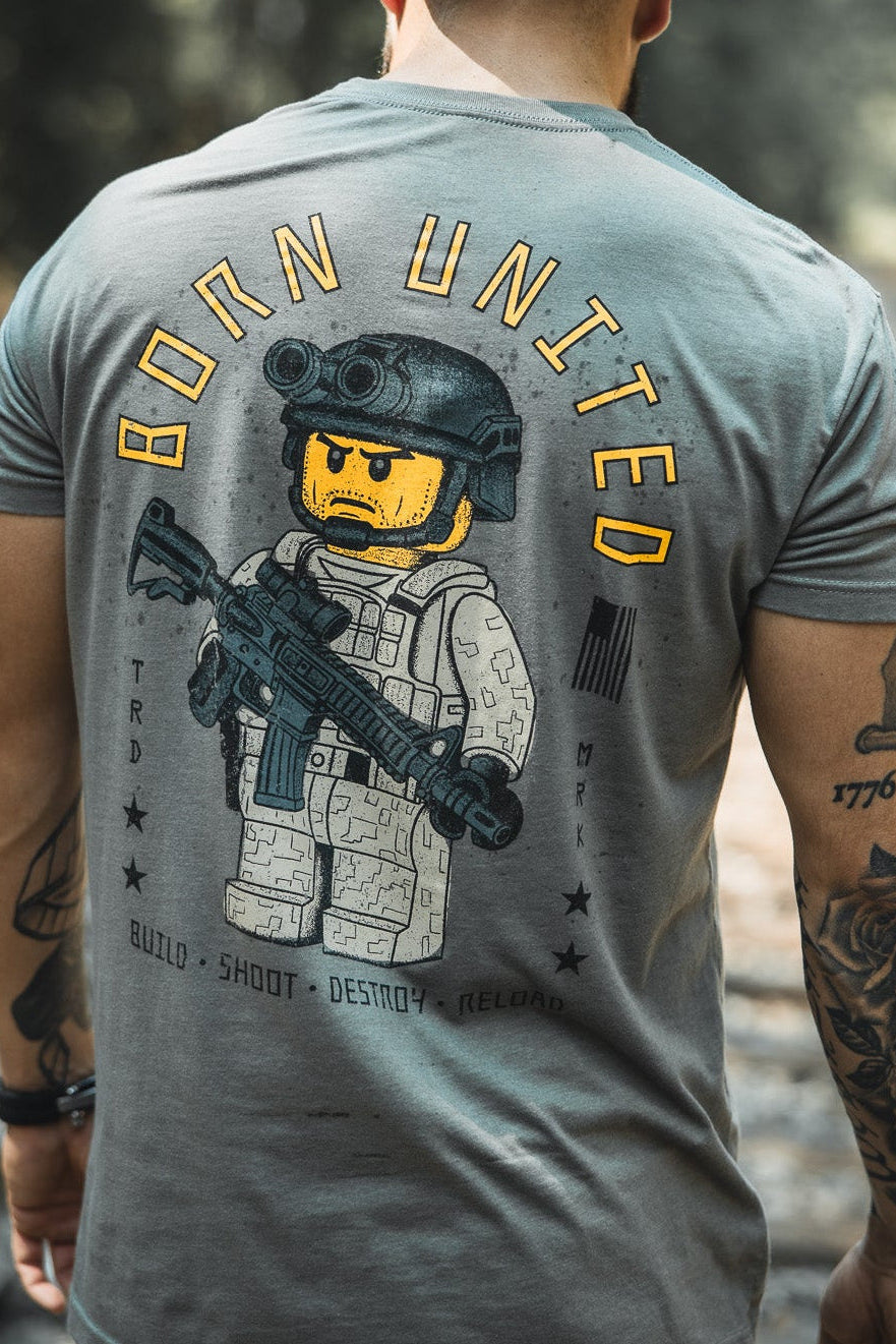 A tattooed man stands on railroad tracks in the Born United Private Label Brick Operator Tee, featuring bold yellow lettering and block-style operator art. He completes his look with a black American flag cap.
