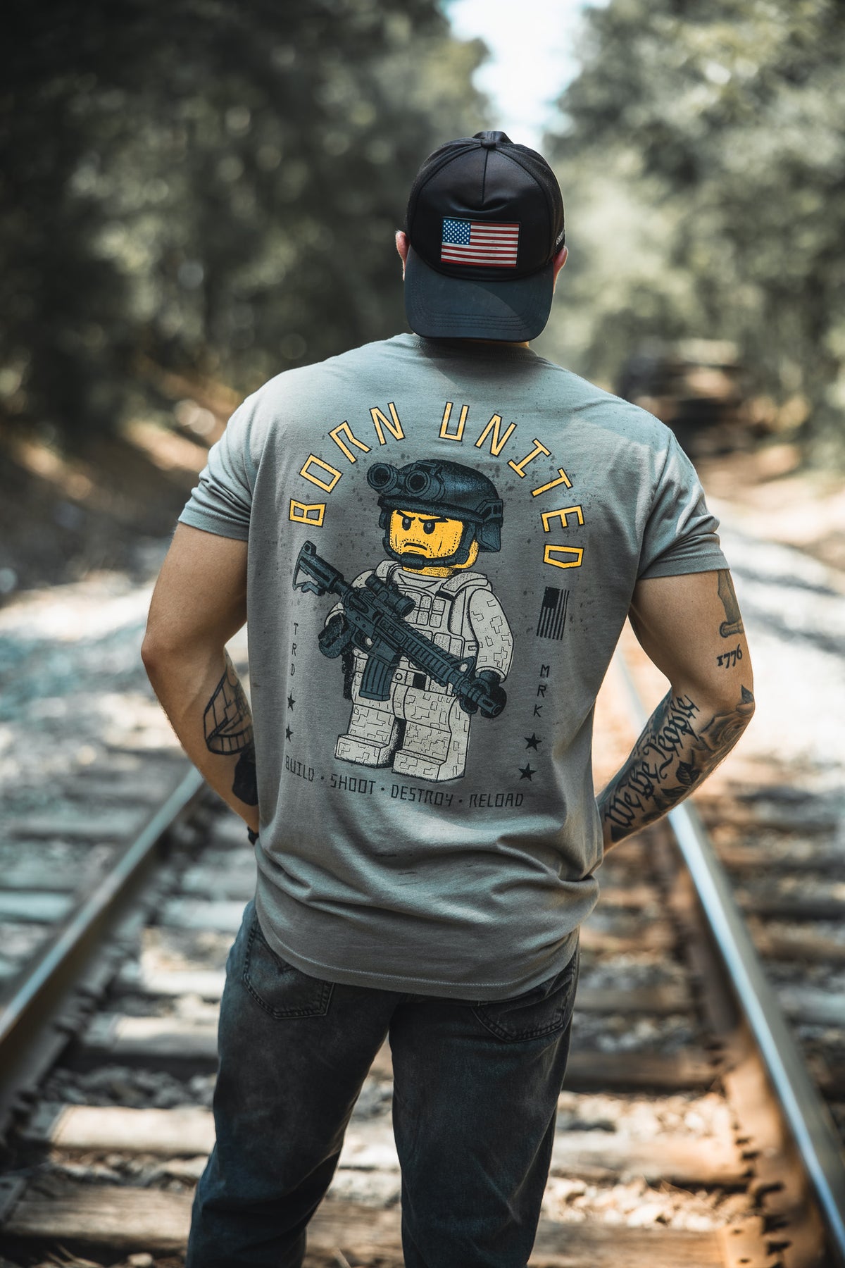 A man stands on train tracks, wearing the Born United Private Label Brick Operator Tee with a LEGO brick soldier graphic and BORN UNITED text, plus an American flag cap and operator-style tattoos. Trees line the railway.