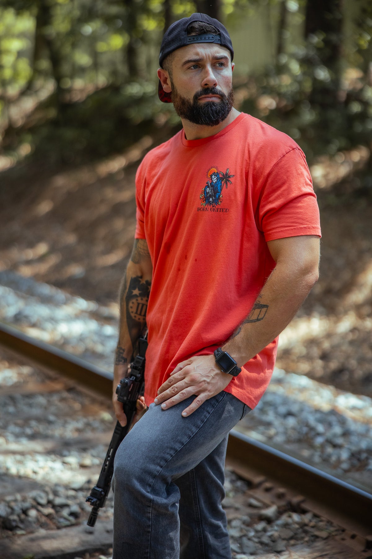 A bearded, tattooed man in the Born United Private Label Always Deadly Tee, jeans, and a backward cap stands by forest railroad tracks holding a rifle, gazing intently into the distance.
