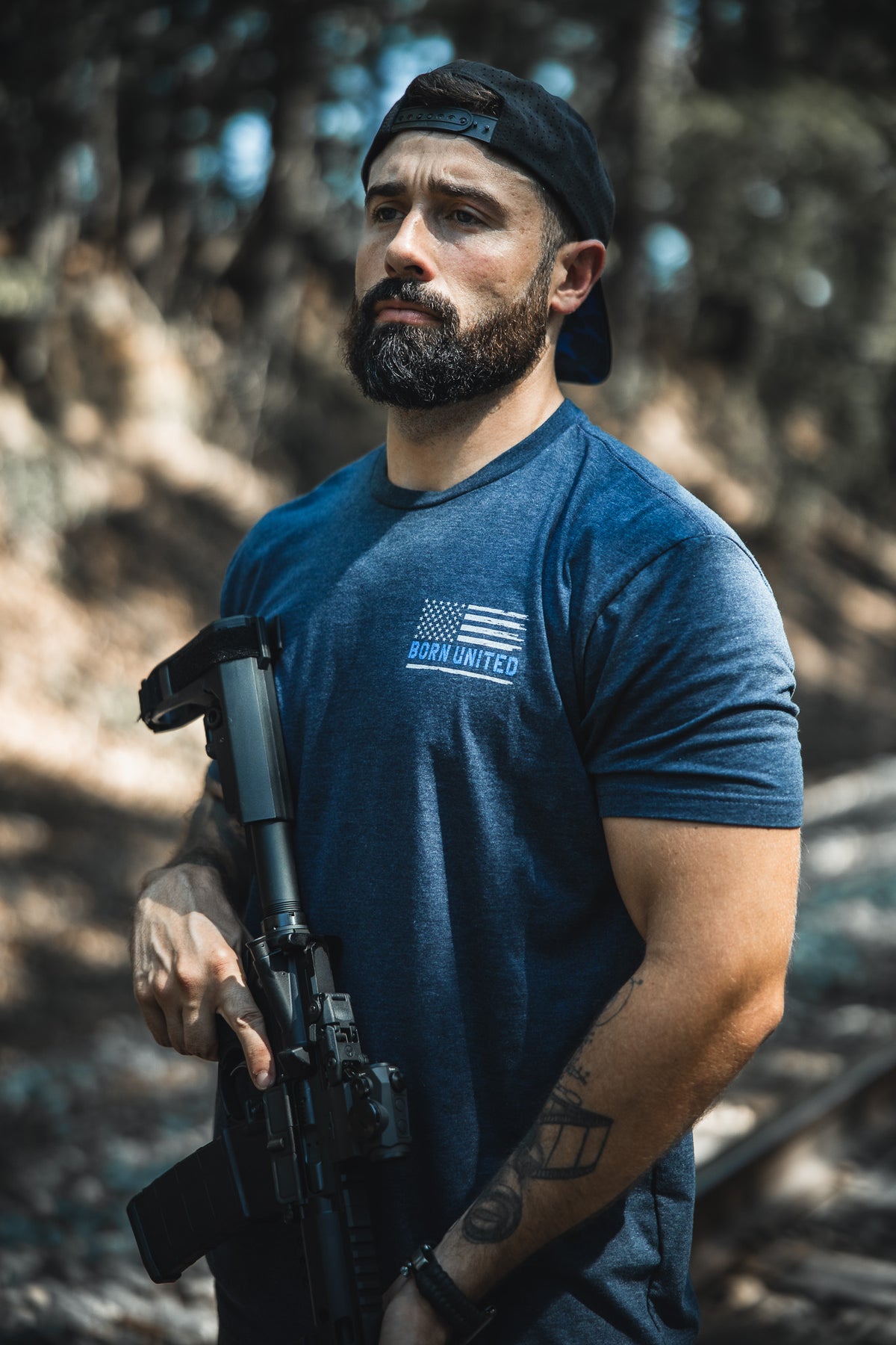 A bearded man in a backwards black cap and a blue The Good Are Unafraid T-shirt by Born United Private Label stands outside, confidently holding a rifle amid trees and sunlight—reflecting bold style and strength.