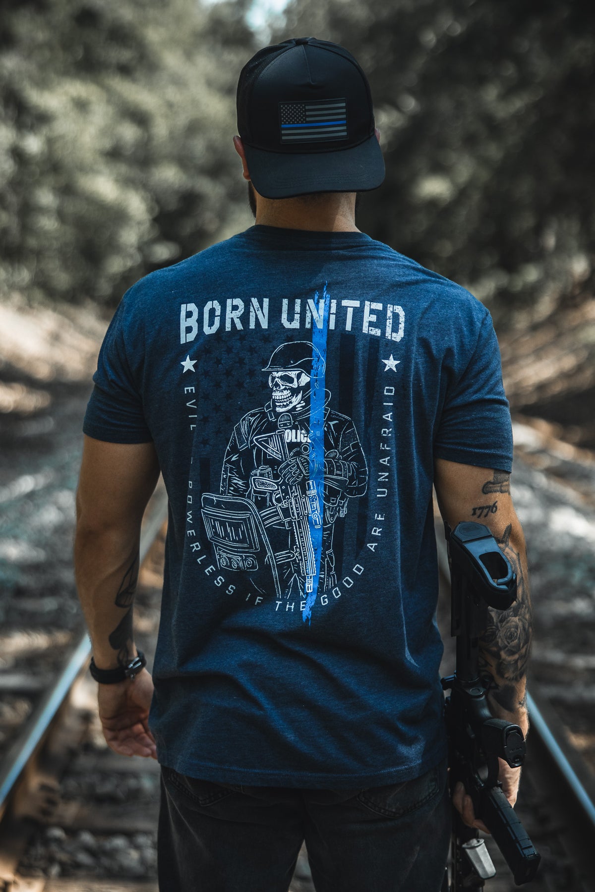 A man stands on railroad tracks wearing a blue The Good Are Unafraid T-shirt by Born United Private Label, featuring a police graphic and blue line. Holding a rifle and wearing an American flag patch cap, he supports Stand Behind the Blue.