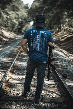 A man stands on train tracks, facing away, wearing Born United Private Label’s “The Good Are Unafraid” T-shirt, jeans, and a backwards cap. He holds a rifle in his right hand, with trees lining both sides of the tracks.