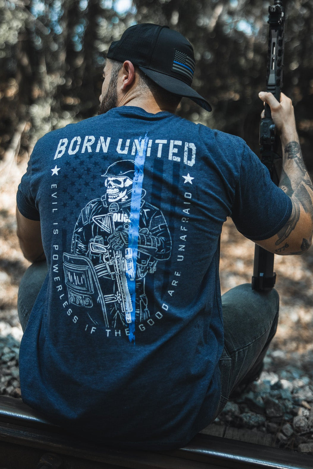 A man wearing the “The Good Are Unafraid” tee by Born United Private Label sits on train tracks in a wooded area, holding a rifle and facing away, embodying the shirt’s message: “Evil is powerless if the good are unafraid.”.