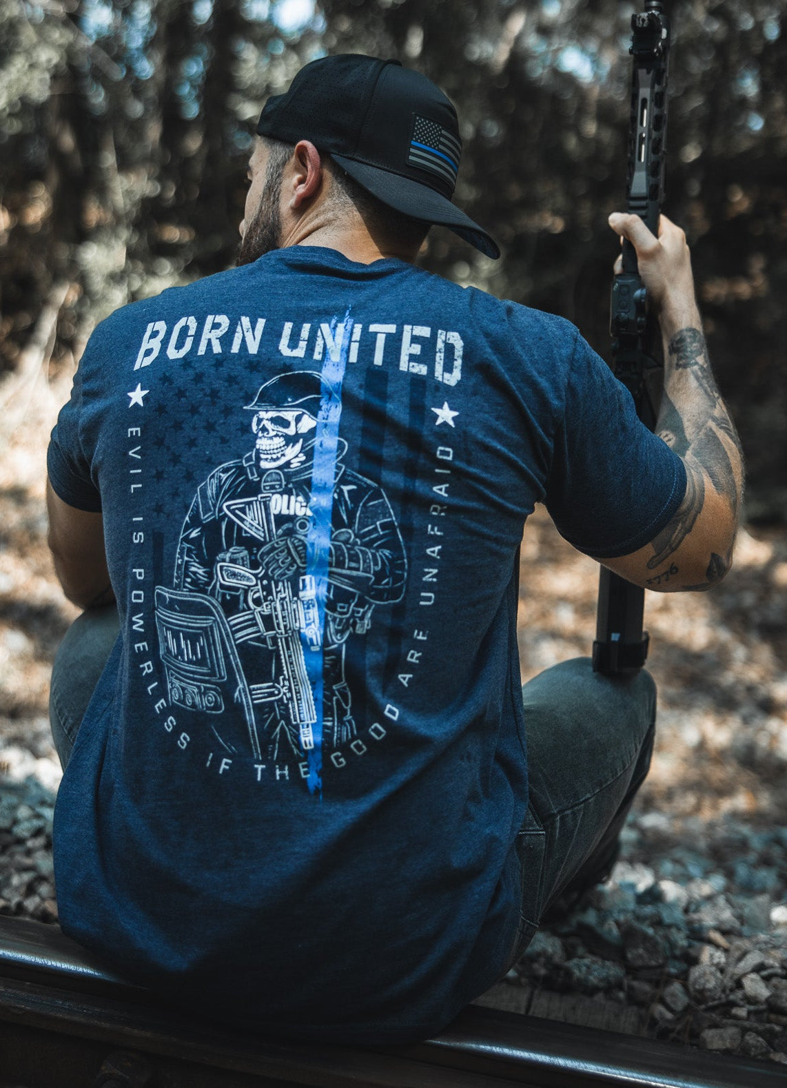 A man wearing the “The Good Are Unafraid” tee by Born United Private Label sits on train tracks in a wooded area, holding a rifle and facing away, embodying the shirt’s message: “Evil is powerless if the good are unafraid.”.