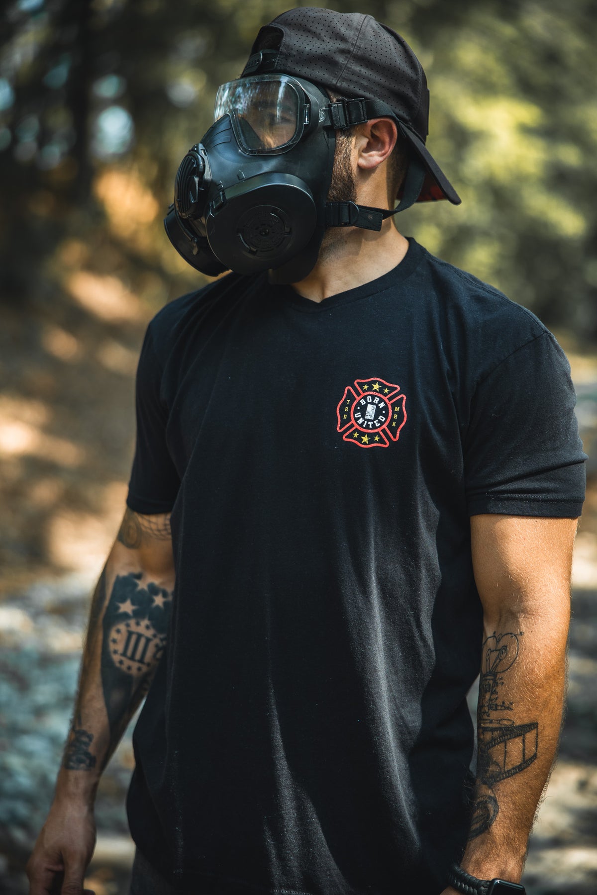 A tattooed individual stands outdoors in a Born United Private Label Brotherhood Forged In Flames black T-shirt, black cap worn backwards, and a gas mask—symbolizing firefighting safety.