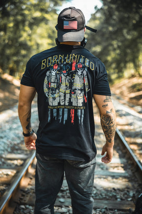 A man stands on railroad tracks, wearing a Born United Private Label black cap with a U.S. flag patch and the Brotherhood Forged In Flames shirt, featuring three firefighters, BORN UNITED, and an American flag. Trees line the tracks behind him.