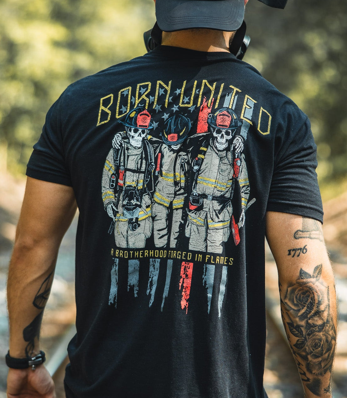A man in a black Born United Private Label Brotherhood Forged In Flames shirt, showing three firefighters and “BORN UNITED,” and a U.S. flag patch cap stands on train tracks in the woods, his tattooed arm visible from behind.