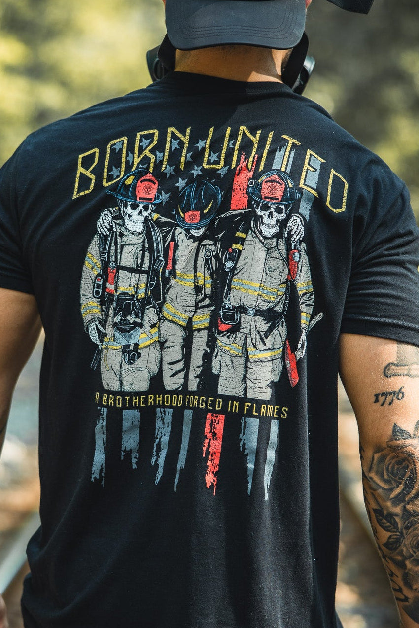 A man in a black Born United Private Label Brotherhood Forged In Flames shirt, showing three firefighters and “BORN UNITED,” and a U.S. flag patch cap stands on train tracks in the woods, his tattooed arm visible from behind.