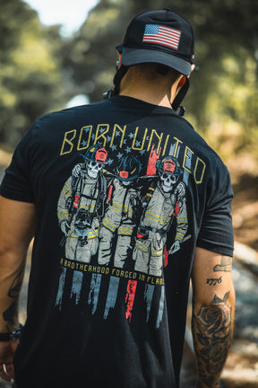 A man wearing a Born United Private Label Brotherhood Forged In Flames black cap with an American flag patch and matching T-shirt featuring three skeleton firefighters, celebrating unity in firefighting.