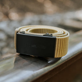 The BU Logo Belt - Coyote by Born United Private Label, featuring an adjustable tan strap and black buckle, is rolled up and positioned on a metal railway track with a blurred outdoor background of trees and sunlight.