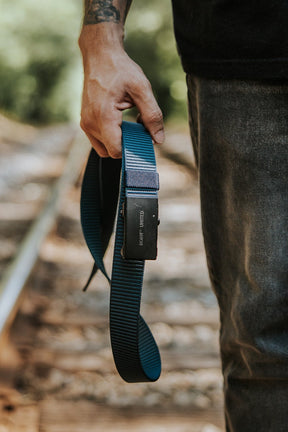 A person with a tattooed arm stands on railroad tracks, holding a Nikon camera strap. They're wearing jeans with the BU Logo Belt - Navy by Born United Private Label and a black shirt, outdoors with green scenery in the background.