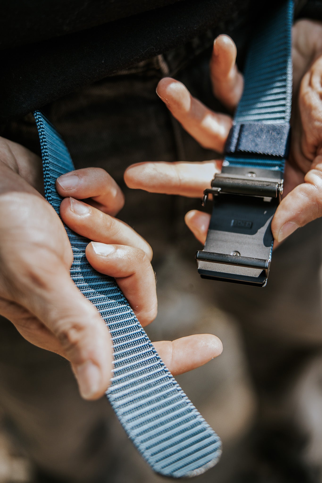 Close-up of hands fastening the BU Logo Belt - Navy from Born United Private Label, featuring an adjustable blue and gray striped nylon strap with a black metal buckle. The blurred background emphasizes the details of the belt and hands.