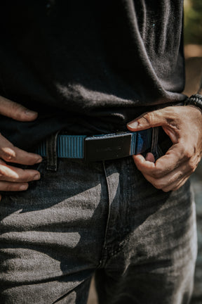 A person wearing a black shirt and dark jeans adjusts the BU Logo Belt - Navy by Born United Private Label, focusing on their hands and waist in natural outdoor lighting.