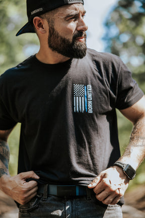 A bearded man outdoors in sunlight, wearing a black “Born United” tee with an American flag print, adjusts his Born United Private Label BU Logo Belt - Navy. He has tattooed arms, a smartwatch, and wears his black cap backwards.