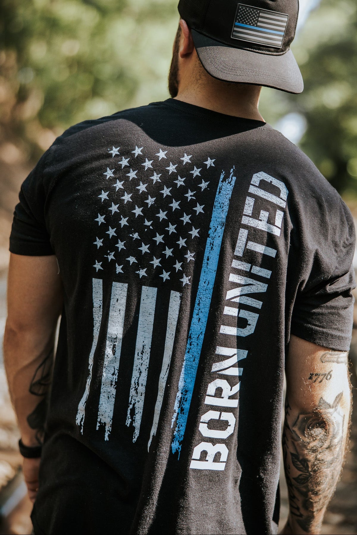 A man stands outdoors on a sunlit path, wearing the Born United Private Label Blue Line Flag Tee—a black T-shirt featuring a blue and white American flag and showing support for Back the Blue.