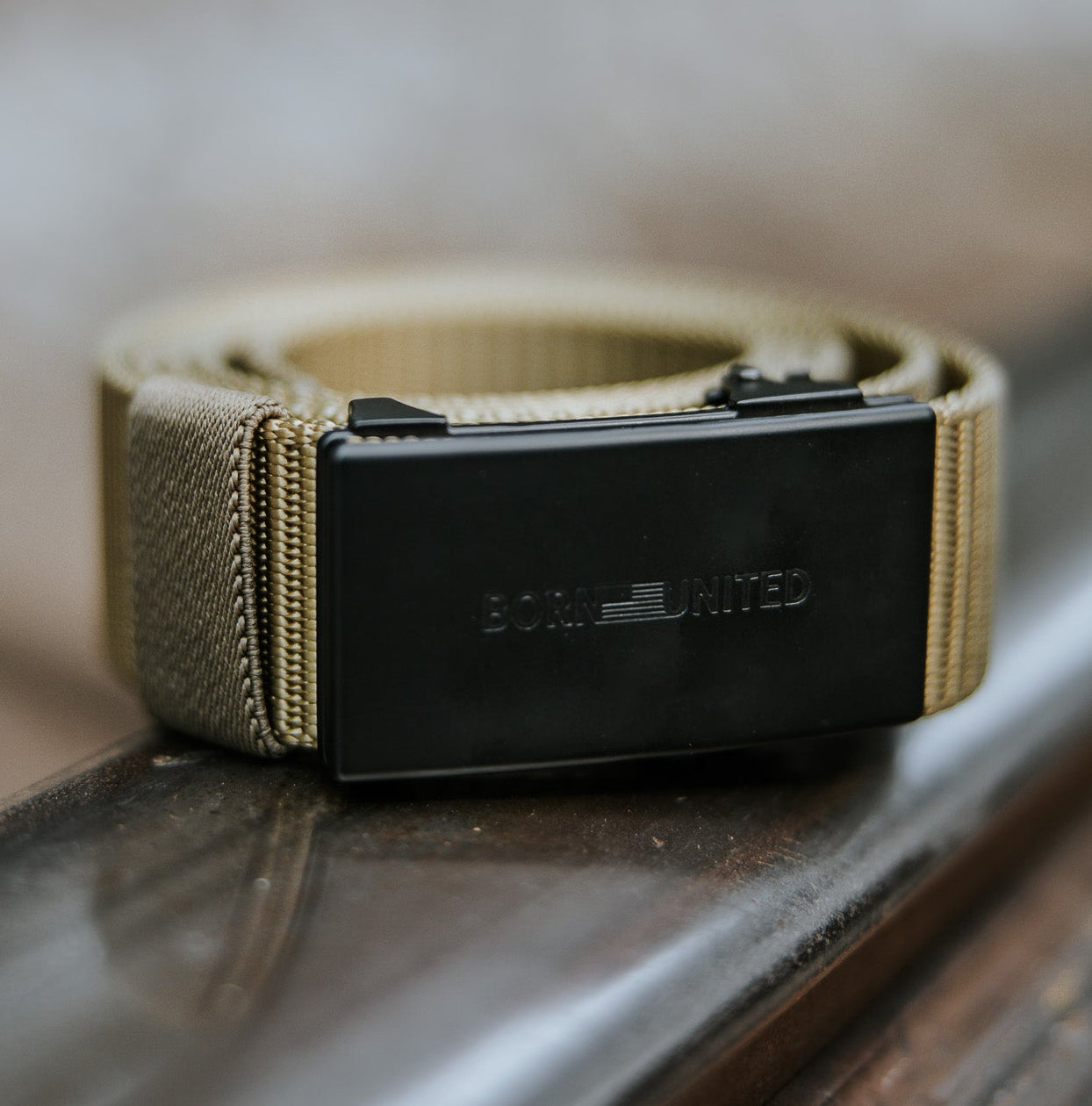 The BU Logo Belt - Coyote by Born United Private Label, featuring a black rectangular buckle engraved with "BORN UNITED," is displayed on a metal railing against a softly blurred background.