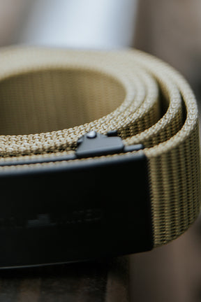 Close-up of the BU Logo Belt - Coyote by Born United Private Label, showing its coiled tan woven strap and black buckle on a surface, with the belt's texture and part of the buckle in focus against a blurred background.