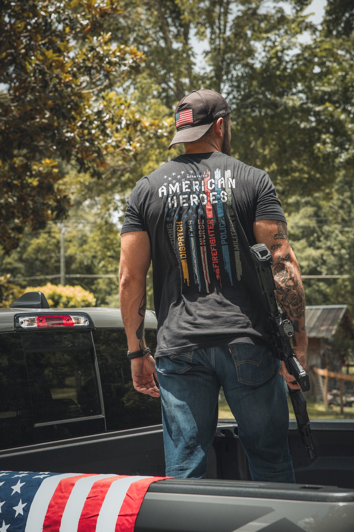 A man stands in the back of a pickup, wearing a Born United Private Label American Heroes Tee with colored lines and names on the back. He holds a rifle, has an American flag patch on his cap, and an American Heroes Flag draped nearby.