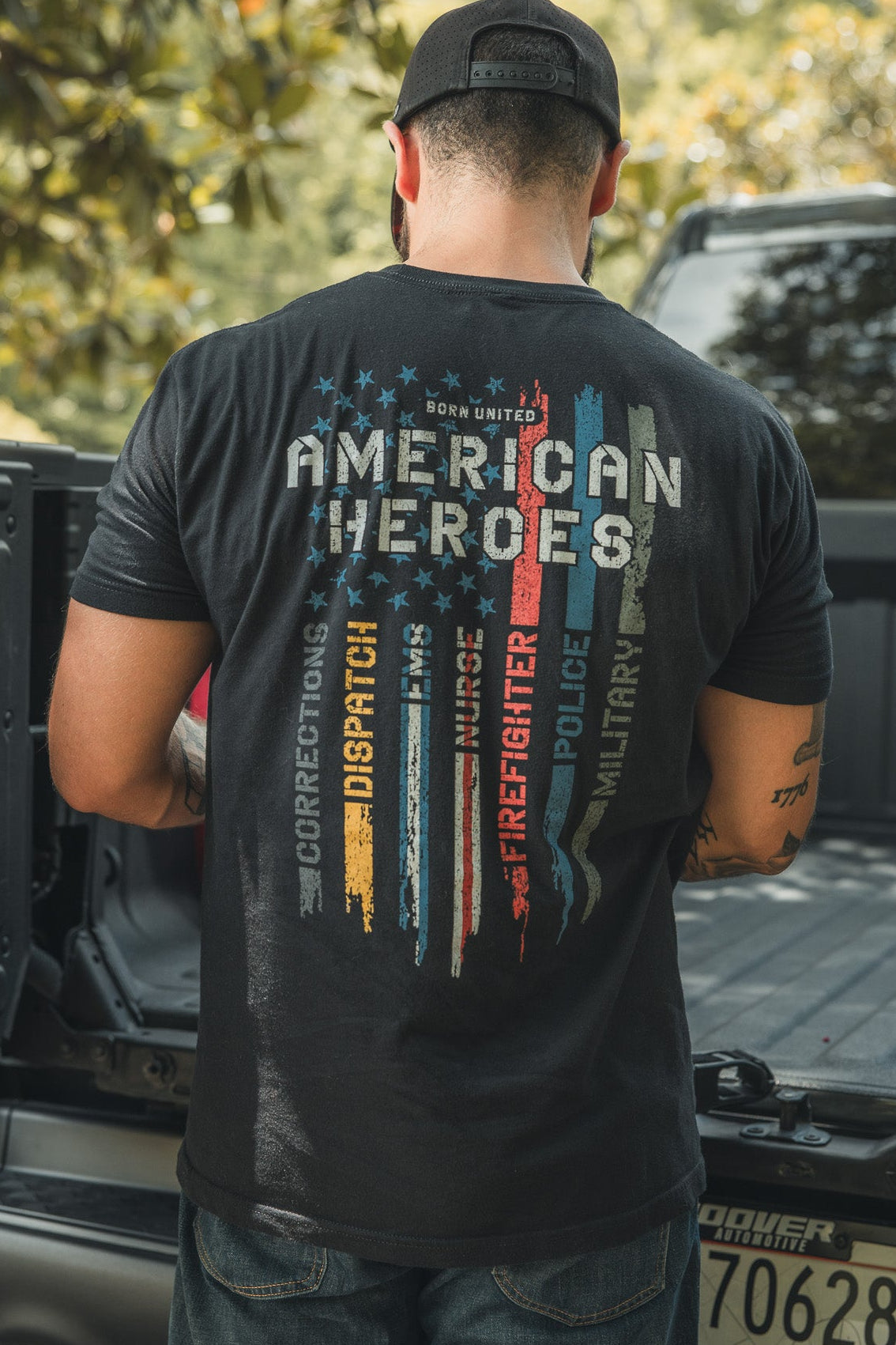 A man stands at the back of a pickup truck, wearing a black cap and an American Heroes Tee by Born United Private Label, featuring colorful stripes and words like Corrections, Dispatch, Firefighter, Police, and Military.